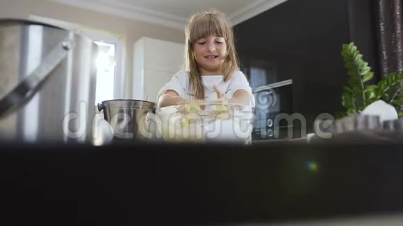 可爱的小女孩留着长发穿着白色连衣裙在家里的现代厨房里用玻璃碗揉面团孩子的手视频的预览图