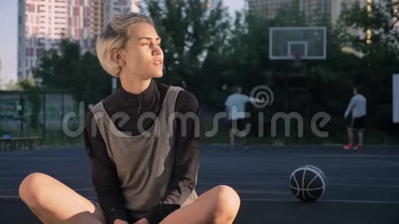 漂亮的金发女人坐在篮球场上对着镜头微笑男人在后台打球女球员视频的预览图