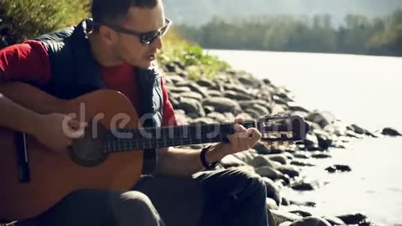 阳光明媚的日子里戴着太阳镜的年轻人在河边慢动作地坐着吉他演奏视频的预览图