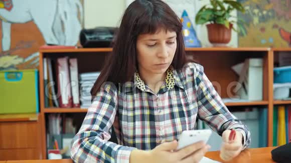 年轻的女老师坐在桌子旁处理文件和智能手机视频的预览图