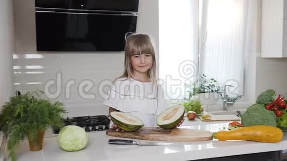那个穿着白色连衣裙的可爱小女孩坐在木板上在家里的厨房里拿着一个绿色的甜瓜切成两半健康食品视频的预览图