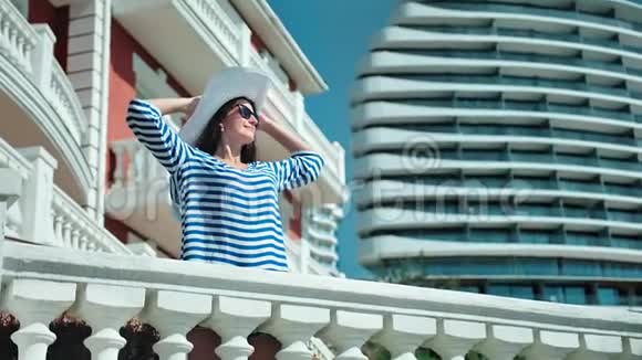 戴着帽子戴着墨镜的优雅女人举起双手享受阳光站在度假阳台上视频的预览图