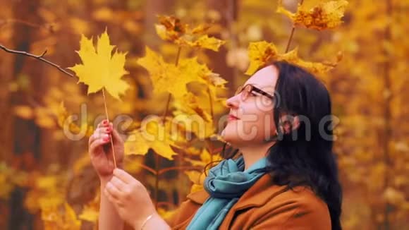 秋天公园里一个戴着眼镜的年轻微笑的女人手里拿着一片花纹的枫叶检查着它视频的预览图