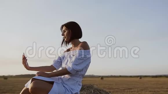 一个漂亮的女孩坐在干草堆上的田野里用电话自拍穿着蓝色裙子的漂亮年轻女子视频的预览图
