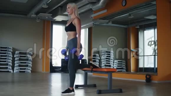 性感金发女人在健身俱乐部锻炼单腿仰卧起坐手持蓝色哑铃侧视图视频的预览图