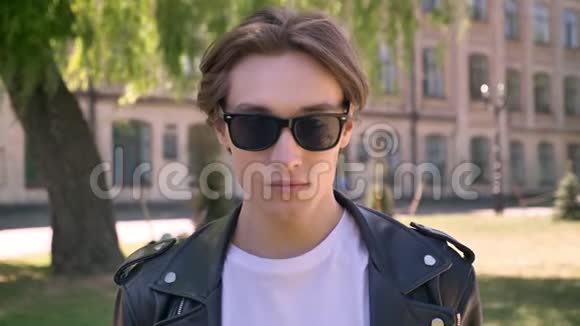 身穿黑色皮夹克的年轻帅哥摘下眼镜看镜头街头背景视频的预览图