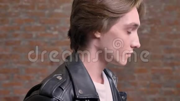 穿着黑色皮夹克的年轻帅哥变成了相机摸着他的头发砖背景视频的预览图