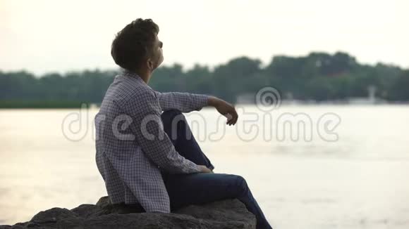 忧郁的男少年独自坐在河边的石头上思考着生活视频的预览图