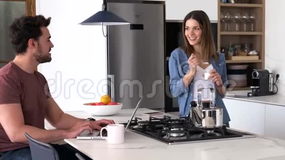 英俊的年轻人用他的笔记本电脑和她的女朋友吃酸奶而站在厨房在家里视频的预览图