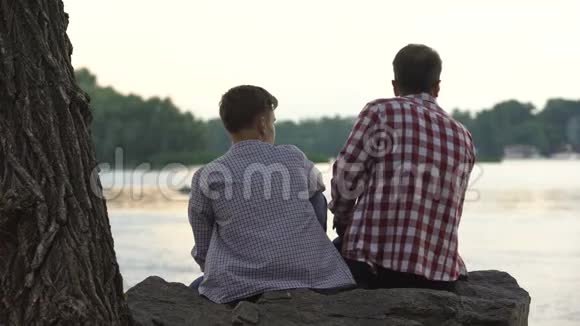 男青少年和他的父亲坐在湖边的石头上谈论生活视频的预览图
