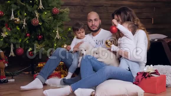 圣诞节父母抱着孩子和小狗年轻的爸爸妈妈看着背景上的摄像机照亮了圣诞树视频的预览图