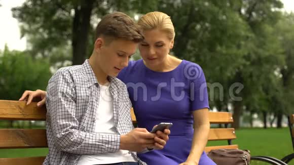 儿子和母亲坐在长凳上看着走路时拍的智能手机照片视频的预览图