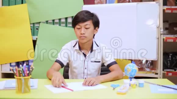 一个男孩坐在桌子旁在笔记本上写东西扔了一支铅笔看着相机视频的预览图