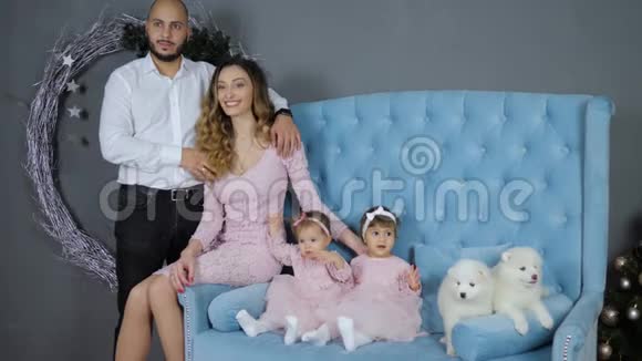 圣诞气氛爸爸带着妈妈和女儿摆姿势和两只小狗在蓝色沙发上合影视频的预览图