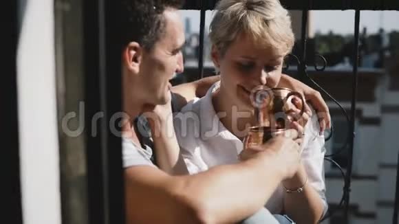 年轻的幸福浪漫夫妇在可爱的公寓阳台上喝咖啡用青铜金属杯子拥抱交谈和微笑视频的预览图