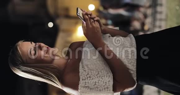 垂直视频迷人的年轻女性使用智能手机站在夜城的街道上对这条了愉快的反应视频的预览图