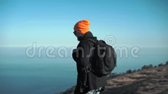背包戴帽子戴太阳镜站在山顶边缘伸展手臂视频的预览图