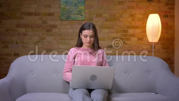 一位年轻漂亮的黑发白种人女性使用笔记本电脑坐在舒适的沙发上微笑的特写镜头视频的预览图