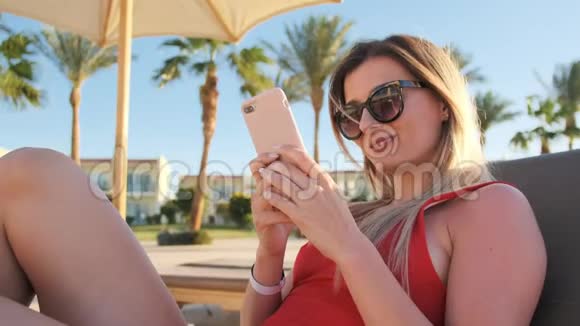 微笑迷人的年轻女孩穿红色比基尼使用她的智能手机在酒店泳池边的肖像假期旅费视频的预览图