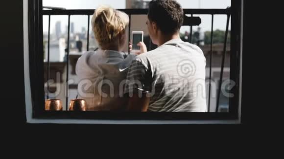 快乐的年轻朋友坐在纽约的一个小阳台上从公寓窗口观看使用智能手机购物应用程序视频的预览图