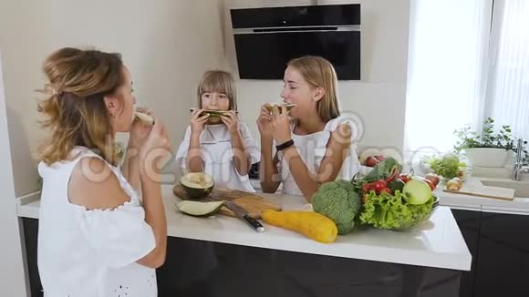 两个可爱的青少年姐妹和一个穿着白色衣服的妈妈在家里的厨房桌子上吃熟瓜健康食品饮食视频的预览图
