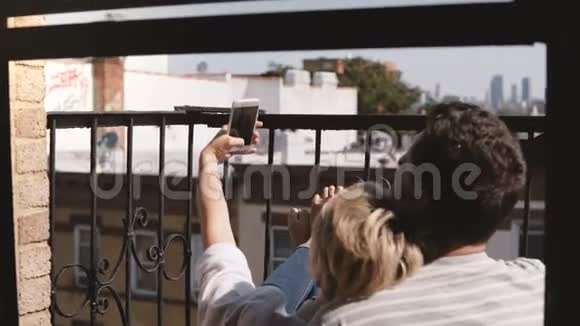 后景幸福浪漫夫妇坐在纽约的一个小公寓阳台上拍智能手机自拍玩得开心视频的预览图