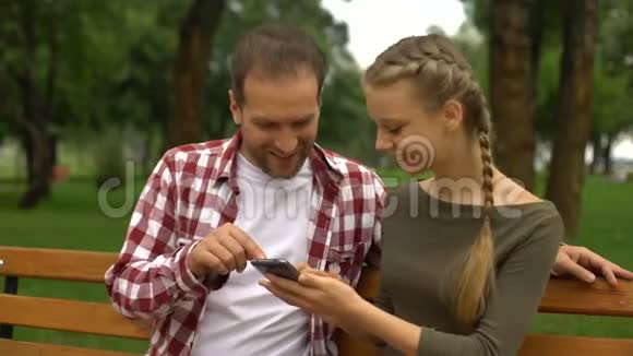 可爱的十几岁的女儿在手机上给父亲播放有趣的视频在公园里放松视频的预览图
