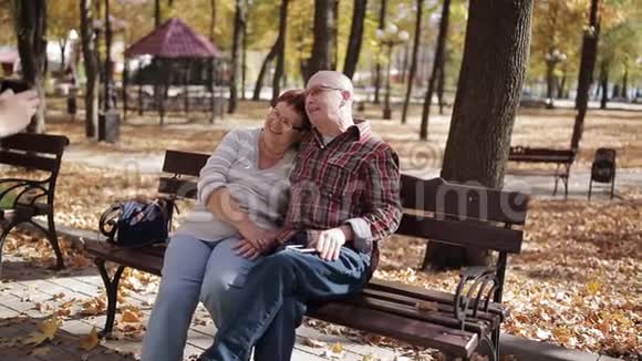 成年儿子在秋天公园坐在长凳上的智能手机上拍摄年迈的父母的照片视频的预览图