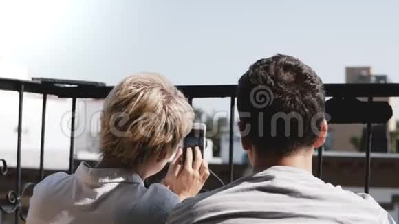 幸福的年轻夫妇坐在阳光明媚的小阳台上在智能手机上拍摄纽约曼哈顿美丽的景色视频的预览图