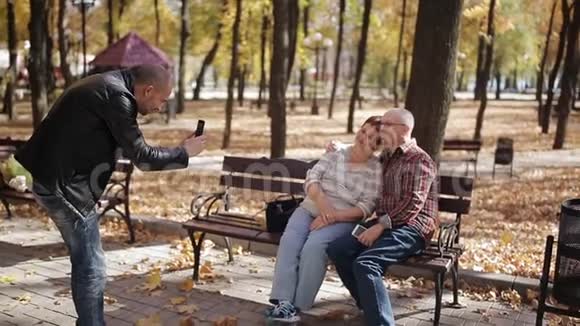 一个成年的儿子和年迈的父母在秋天公园散步给坐在长凳上的父母拍照视频的预览图