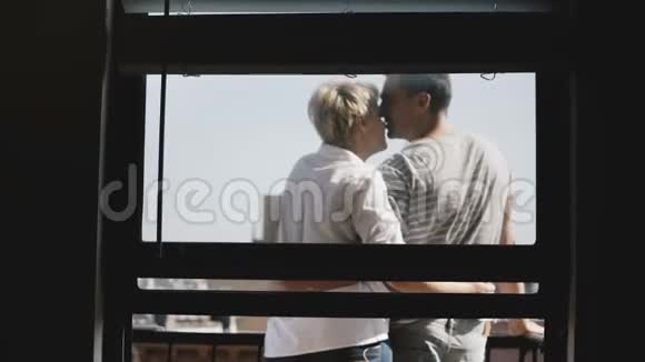 可爱的年轻浪漫夫妇站在一个可爱的小阳光阳台上轻轻地亲吻透过公寓的窗户观看视频的预览图