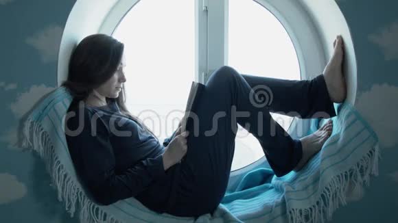 年轻女子正在读一本书一位美丽的年轻姑娘坐在一扇圆窗上看书视频的预览图