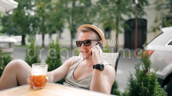 戴着帽子戴着太阳镜的英俊男子正在打电话微笑着他正在喝一杯鸡尾酒放松一下视频的预览图