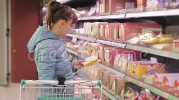 超市里的女人年轻的白种人女人穿着蓝色夹克读着奶酪标签把它放进手推车里视频的预览图