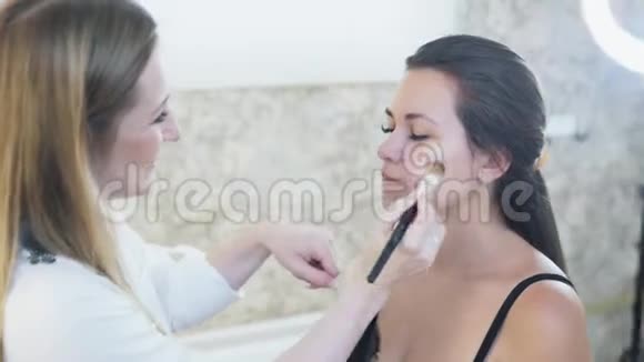 一位年轻的金发碧眼的化妆师用一个遮瑕膏在她脸上画一个年轻的黑发女孩视频的预览图