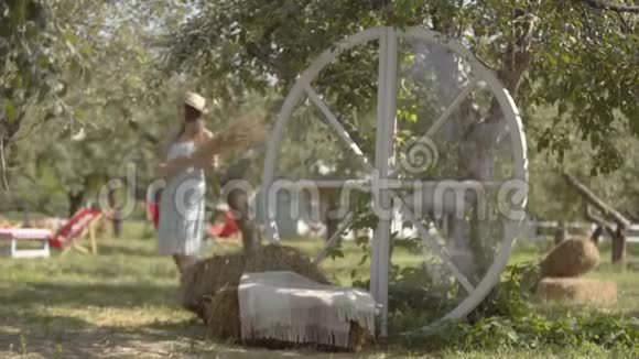 漂亮的年轻女子戴着草帽穿着白色的长裙在绿色的夏日花园里抱着一堆草农村视频的预览图
