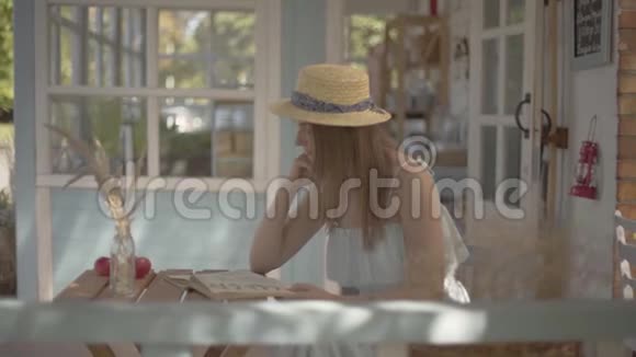 年轻的农村女孩戴着草帽和白色连衣裙坐在户外的小木桌上看书农村视频的预览图