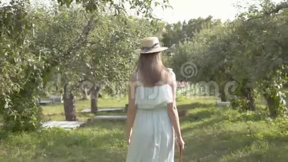漂亮的年轻女子戴着草帽穿着长长的白色连衣裙穿过绿色的夏天花园免费农村生活视频的预览图