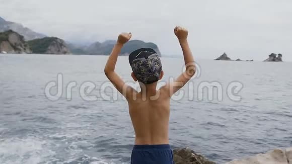 后景七八岁的小男孩站在海边的一块岩石上举起双手为生活感到高兴关于视频的预览图