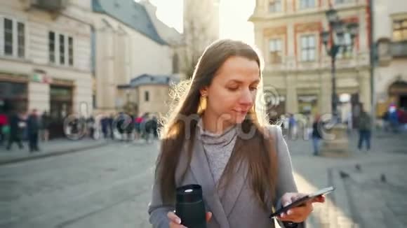 年轻的女人手里拿着一个保温杯在日落时用智能手机沿着一条老街走着交流社会视频的预览图