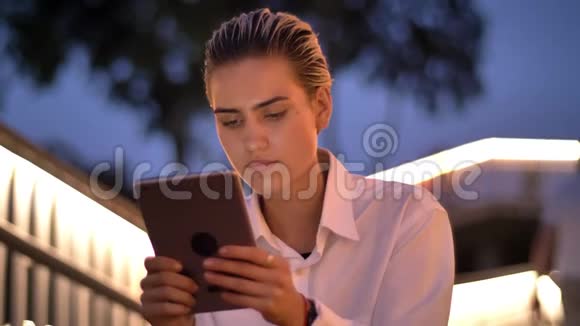 年轻的现代女性坐在梯子上晚上敲着平板电脑微笑着背景上模糊的灯光视频的预览图