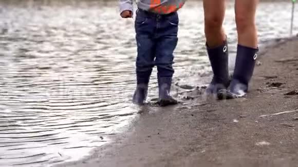 妈妈和孩子穿着橡胶靴在湖边散步在水和潮湿的沙子上行走视频的预览图