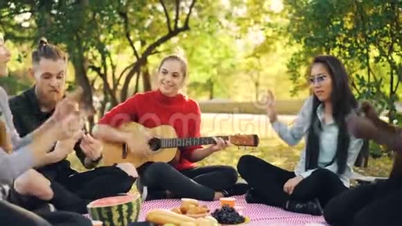 当美丽的女孩在公园野餐时弹吉他时嬉戏的年轻人在唱歌和动手视频的预览图