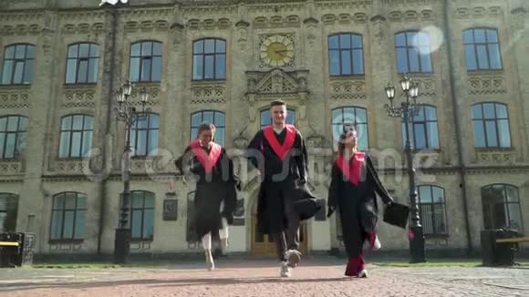 三个穿着黑色和红色壁炉的学生在大学大楼前庆祝他们的毕业新毕业视频的预览图