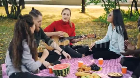 漂亮的年轻女士正在弹吉他而她的朋友正在唱歌听音乐休息在公园的格子呢食物视频的预览图