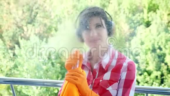 透过窗户观看微笑的女人戴着耳机戴着手套喷洒清洁产品使用清洁窗口视频的预览图