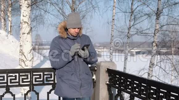 穿着带毛衣罩的蓝羽绒服的人拿起手机在冬季公园里脱下手套视频的预览图
