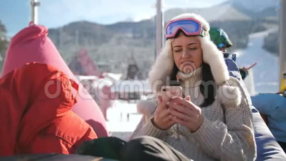 在高山滑雪场放松的年轻女子一个女孩坐在滑雪场的咖啡馆里用智能手机视频的预览图