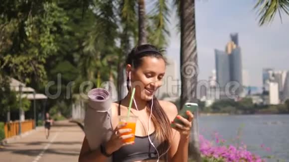 美丽的适合混合种族妇女使用手机应用程序和饮用新鲜橙汁后上午瑜伽练习视频的预览图
