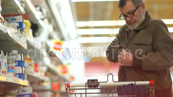 一位留着灰色胡子和眼镜的老人在超市里挑选出一个奶酪然后把它放进手推车里乳制品视频的预览图